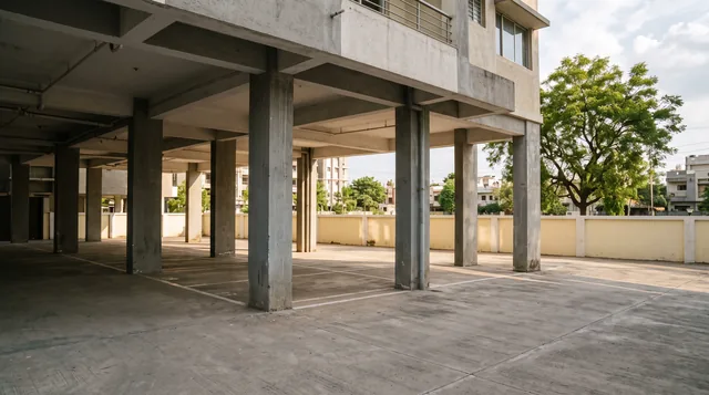 An empty stilt parking level under a mid-rise apartment block, painted bays between square concrete columns, a cream boundary wall and a large tree in afternoon light.