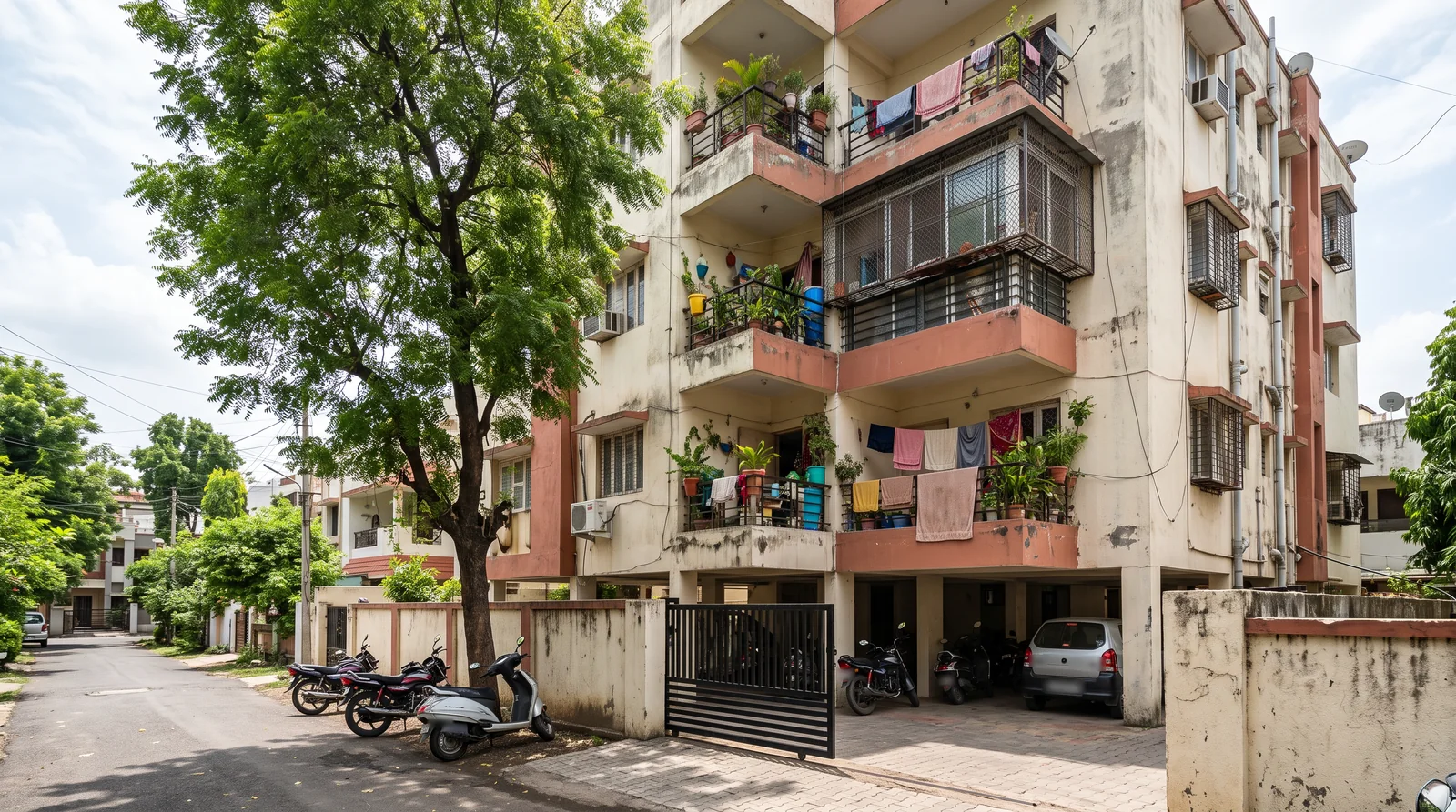 A weathered five-storey apartment building with terracotta balconies full of potted plants and drying clothes, a neem tree in front and two-wheelers parked by its gate.
