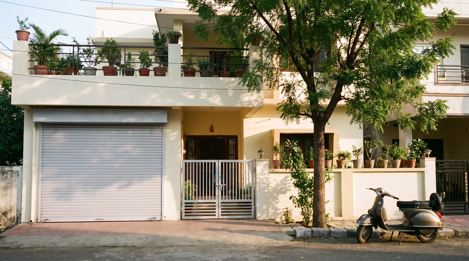 A cream two-storey house in a colony lane with a closed light-grey roller shutter on its ground-floor front room, a white gate beside it, potted plants on the terrace and a scooter under a neem tree.