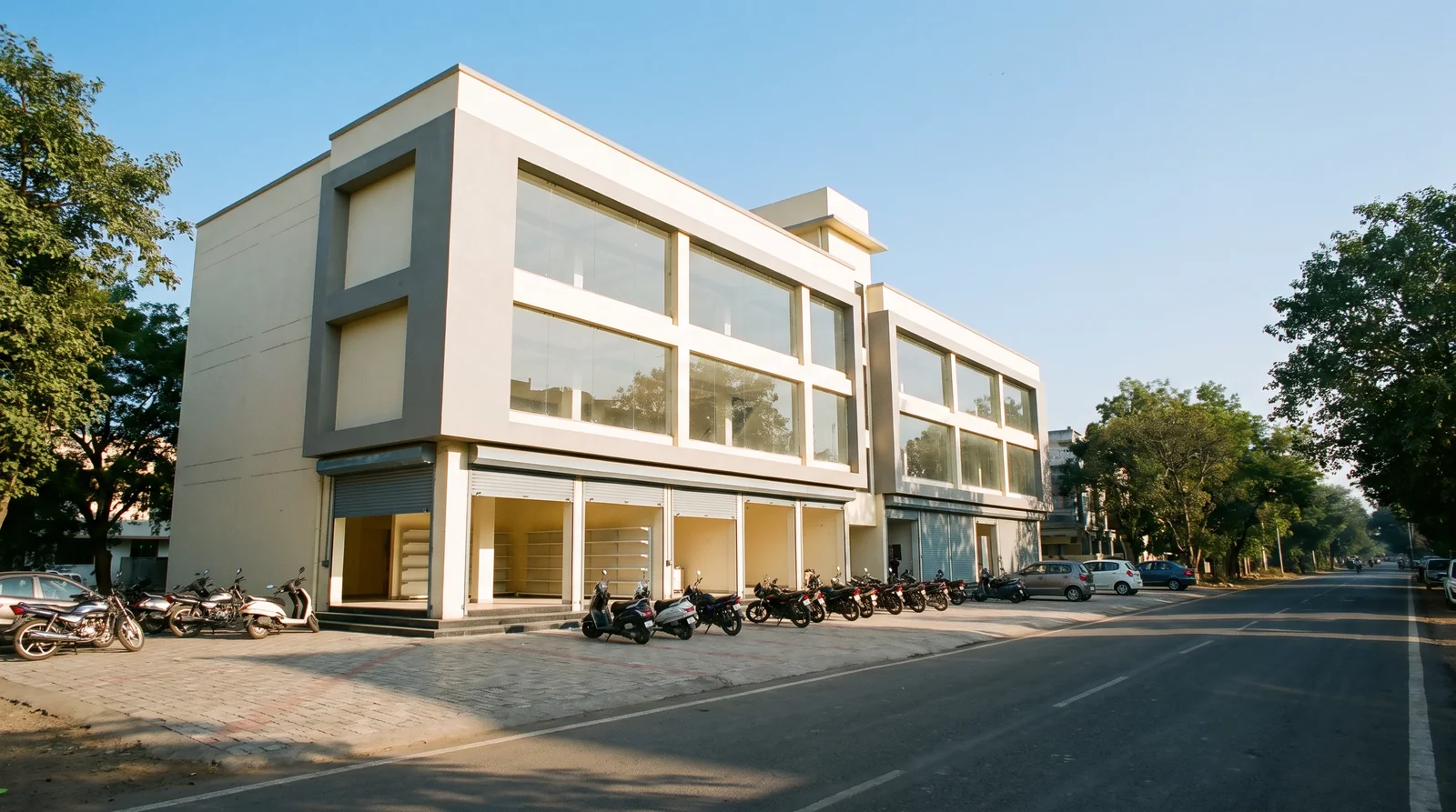 A new three-storey commercial building on a tree-lined road, empty shops with open roller shutters on the ground floor and glass-fronted offices above, two-wheelers parked along its paved front.