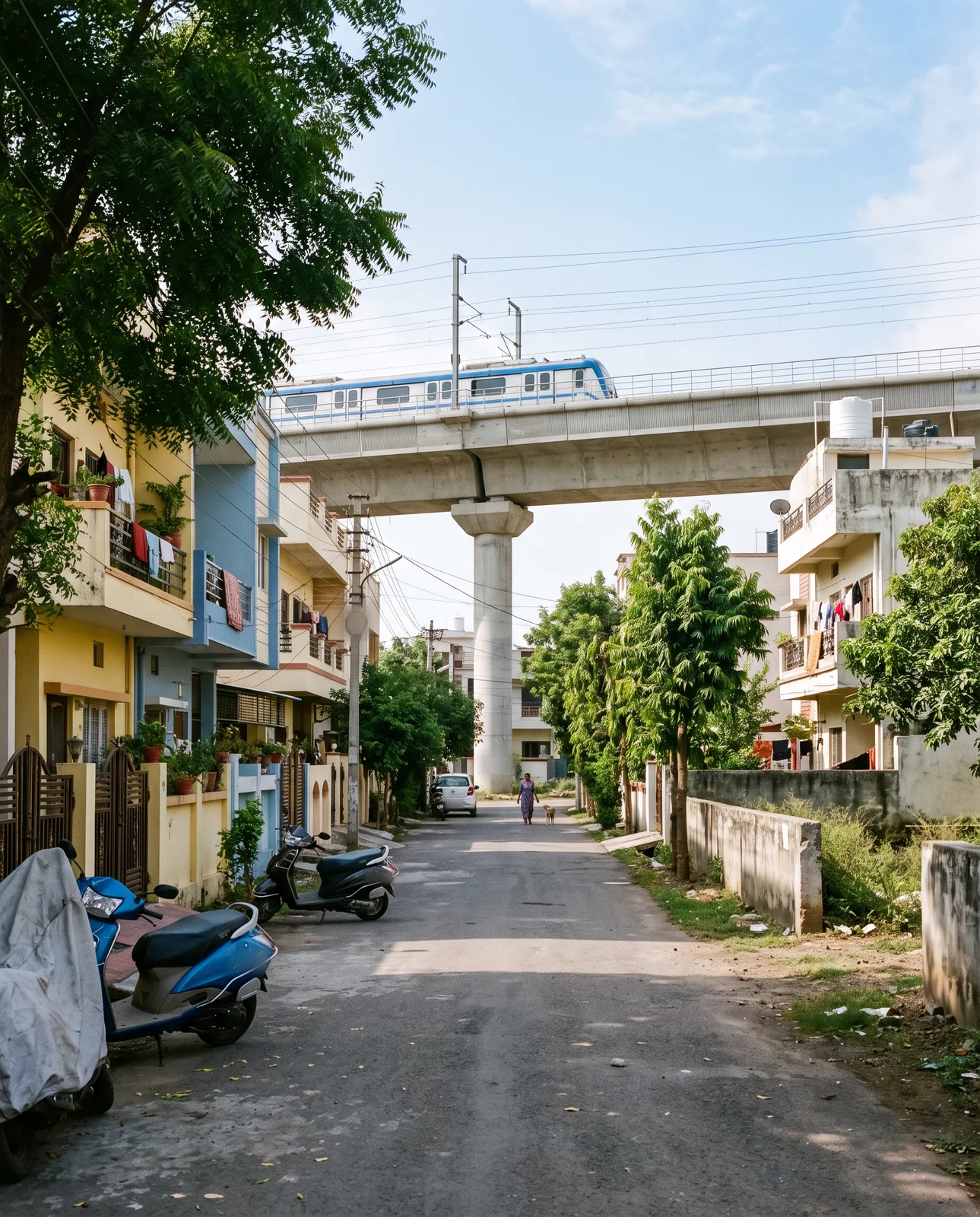 A quiet residential lane with parked scooters and two-storey houses, an elevated metro deck crossing above it on a single pillar.