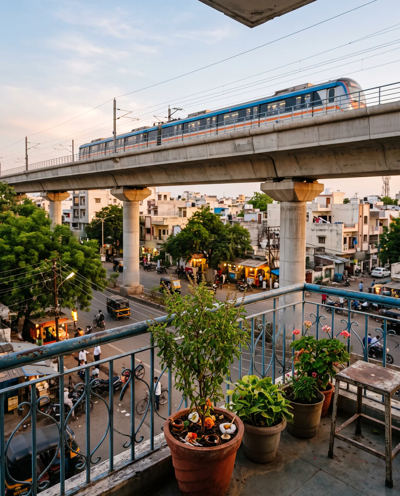 From a first-floor balcony with potted plants and a blue railing, a metro train passes on an elevated viaduct above a busy street at dusk.