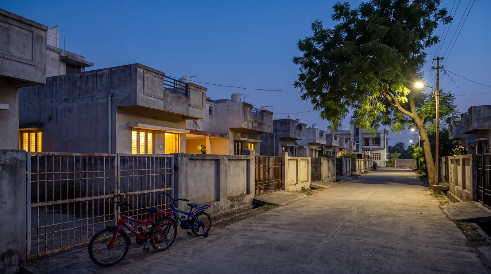 A quiet residential lane at dusk with lit windows, iron gates, two children's bicycles against a wall and a streetlight coming on.