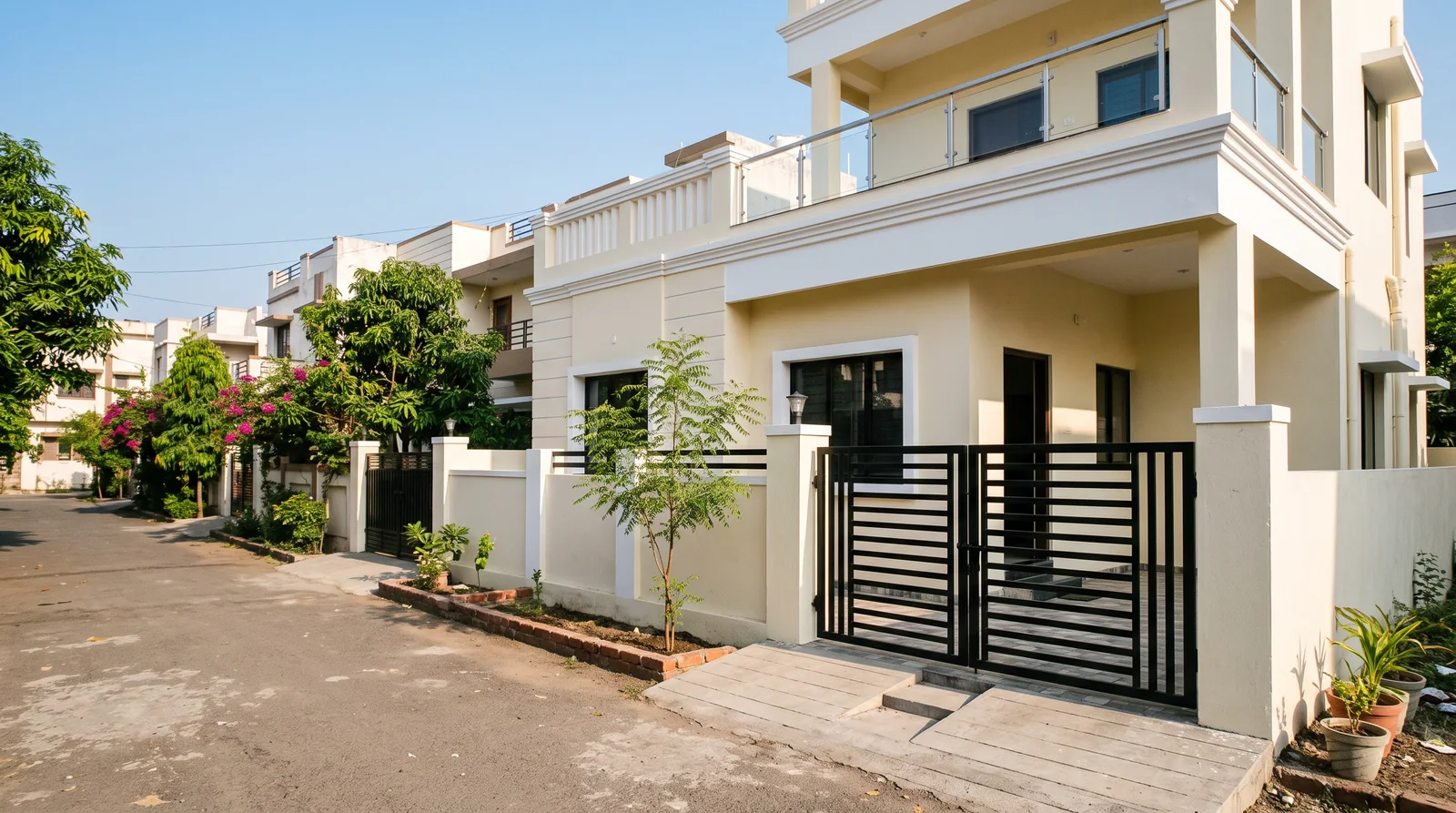 A newly finished cream house with a black slatted gate and a young neem sapling planted along its boundary wall, on a quiet colony street in morning light.
