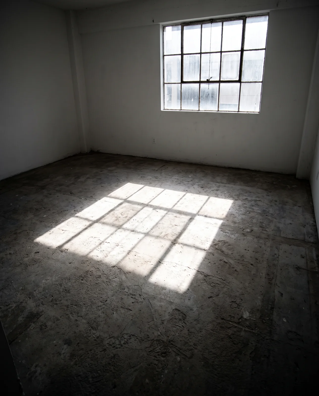 A bare room with a rough concrete floor and white walls, a gridded window throwing a bright patch of light onto the ground.