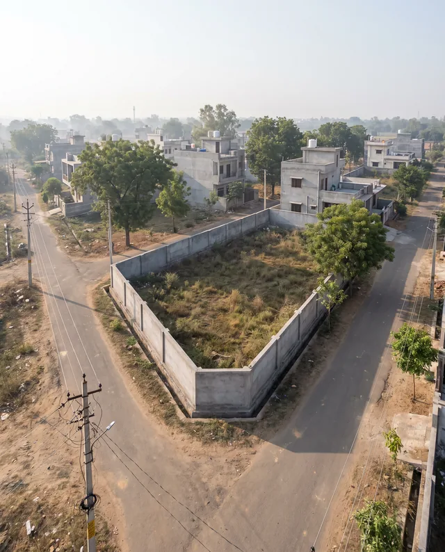 Seen from above, a walled empty corner plot sits at the junction of two roads in a half-built colony, houses and trees behind it