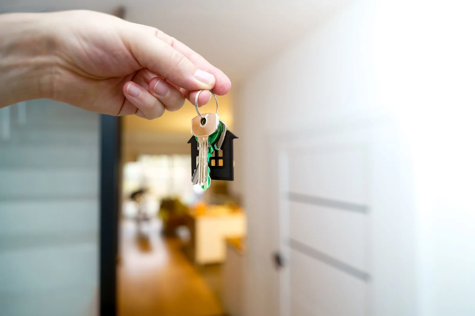 A hand holds up a bunch of keys on a ring with a small black house-shaped key fob, in the hallway of a bright, blurred home.