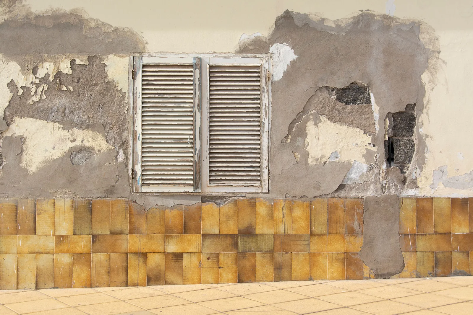 An old wall with plaster flaking off in large patches around a pair of weathered wooden louvred shutters, above a band of worn ochre tiles.