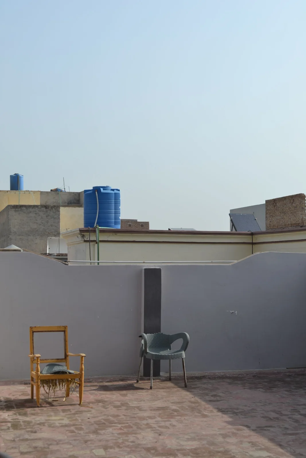 A flat rooftop terrace with a grey parapet wall, two empty chairs, and blue water tanks and a solar panel on the neighbouring roofs.