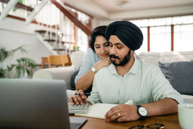 A man in a dark turban works a calculator over a notebook at a low table while his wife rests her chin on his shoulder, a laptop open in front of them.