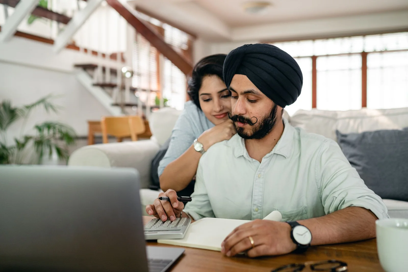 A man in a dark turban works a calculator over a notebook at a low table while his wife rests her chin on his shoulder, a laptop open in front of them.