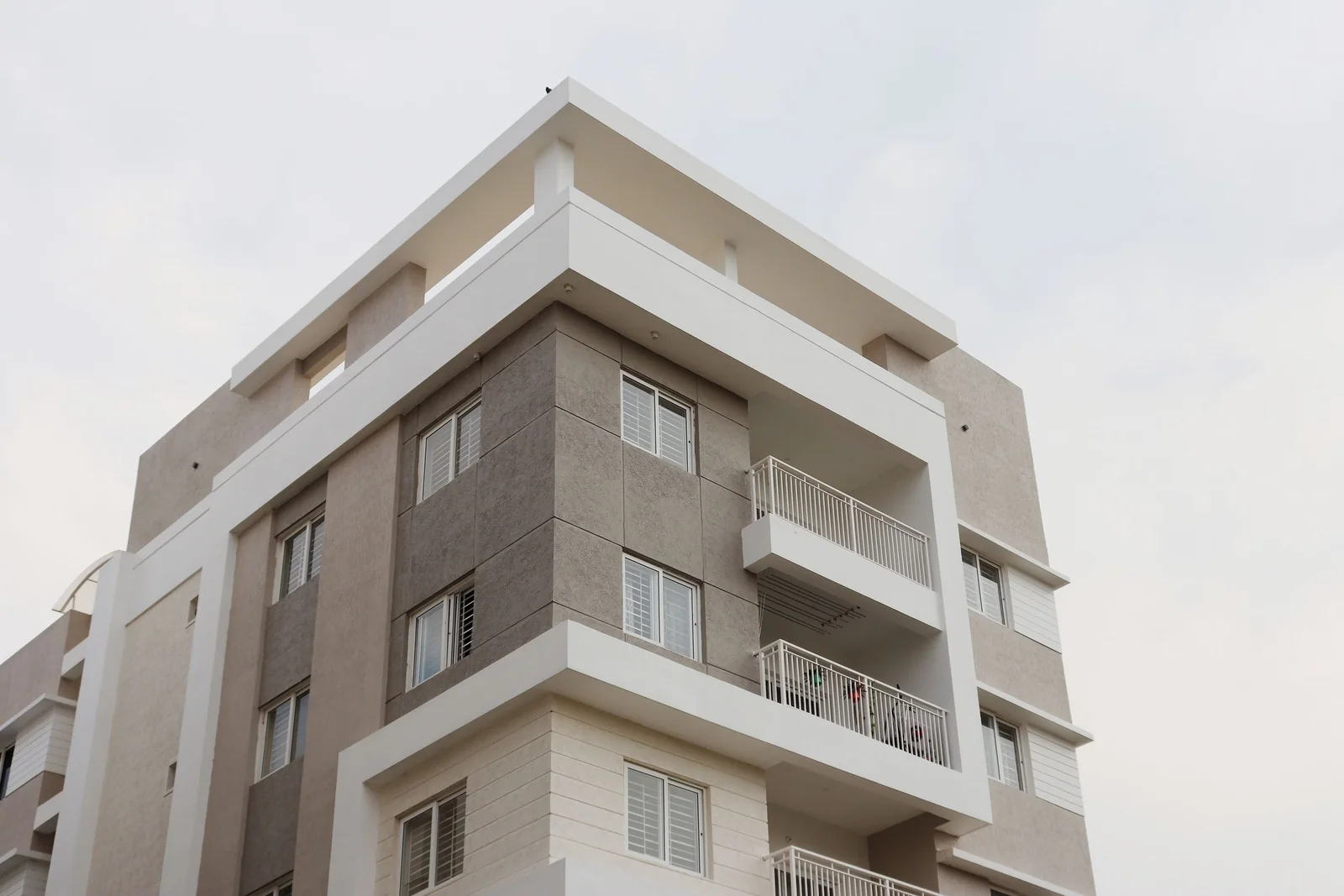 The upper floors of a grey and white apartment building with balconies and shuttered windows against a pale sky.