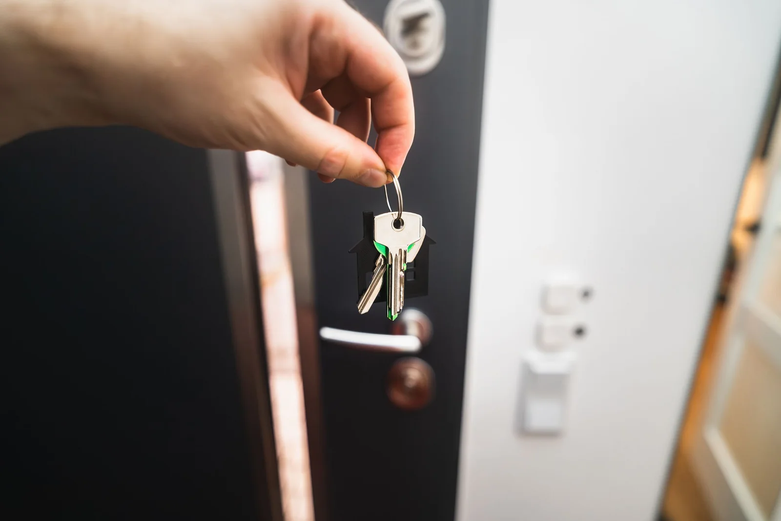A hand holds up a ring of keys with a house-shaped key fob in front of an open dark front door.
