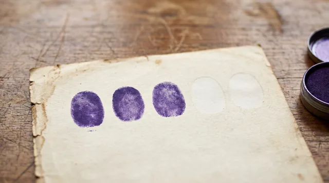 Three purple thumbprints in a row on an old sheet of paper, with two blank ovals beside them and ink pads at the edge.