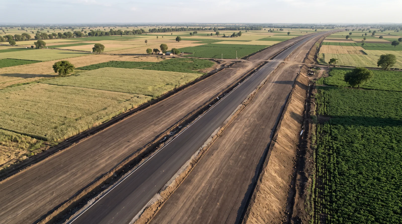 From the air, a very wide new road is being built straight through flat farmland, a fresh asphalt lane in the middle of a broad graded earth corridor, green and golden fields on both sides.