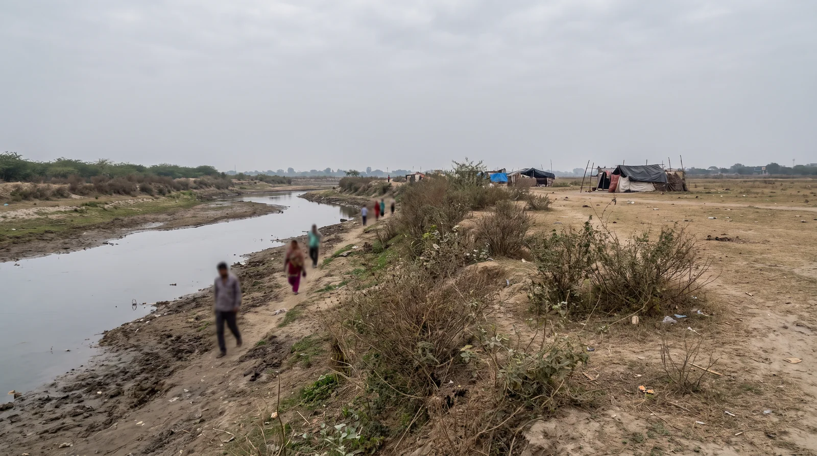 A shallow river channel runs low between muddy banks on an overcast day, a footpath and scrub along one side and makeshift tents in the distance.