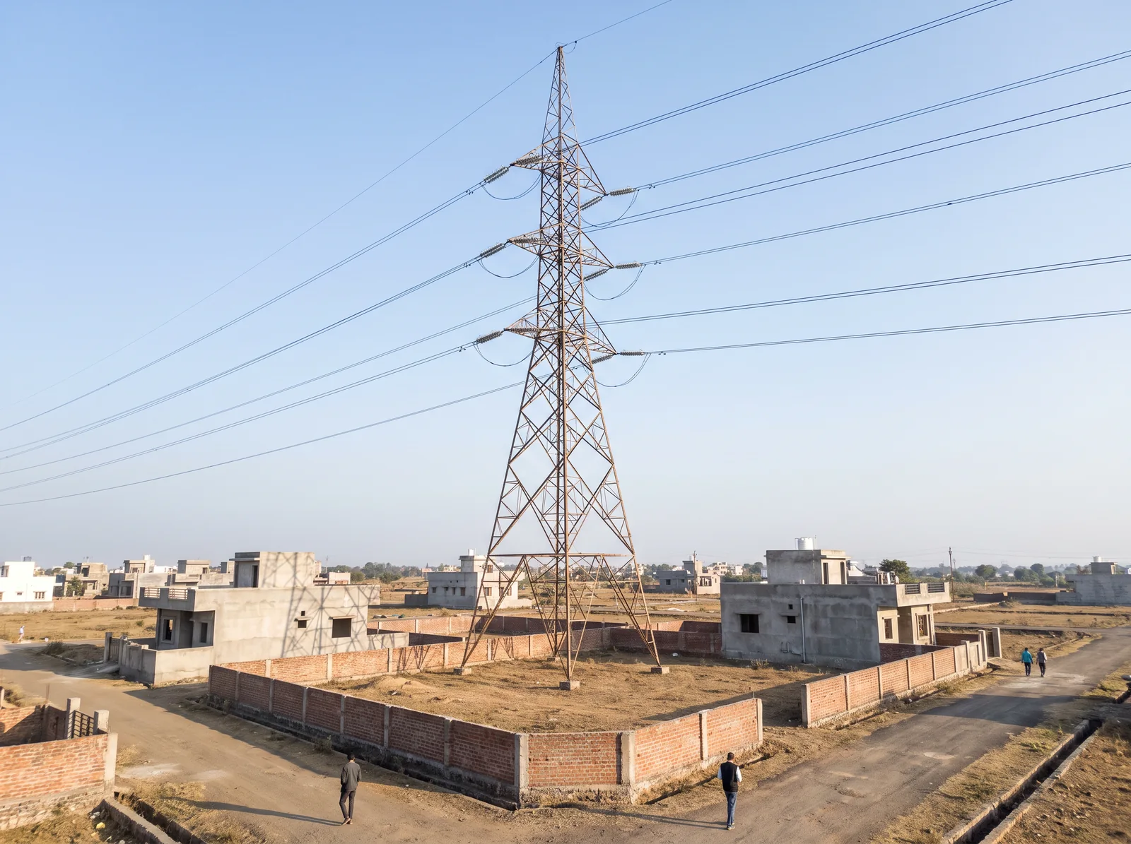 A tall steel transmission tower stands inside a walled empty plot in a new colony, its lines passing over half-built houses on either side.