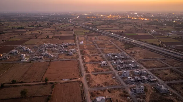 An aerial view at dusk of a plotted colony on the edge of a city, its grid of empty plots and a scatter of grey concrete houses laid out between farm fields, with a lit highway and the city's lights beyond.