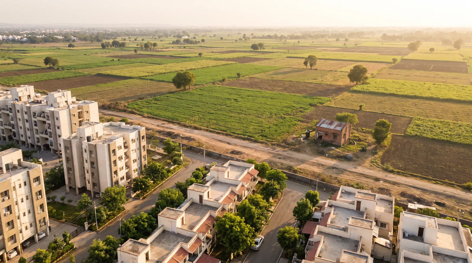 Apartment blocks and flat-roofed houses on tree-lined roads in the foreground give way, across a dirt road, to open green and brown farm fields stretching to the horizon.