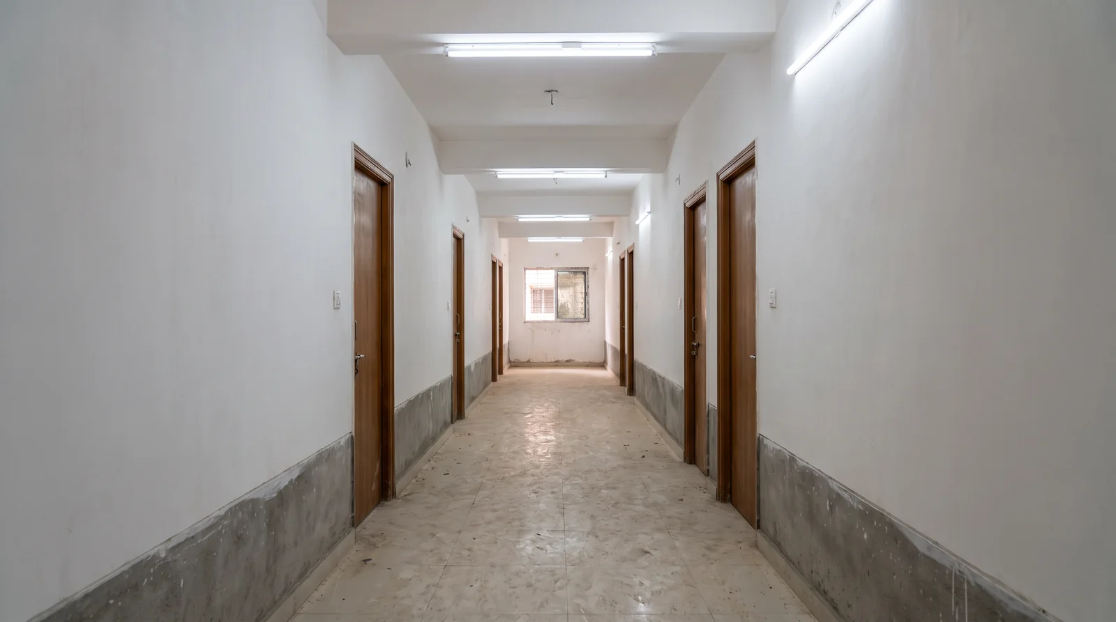 A long, freshly plastered corridor in a new residential block with closed wooden doors on both sides and tube lights along the ceiling.