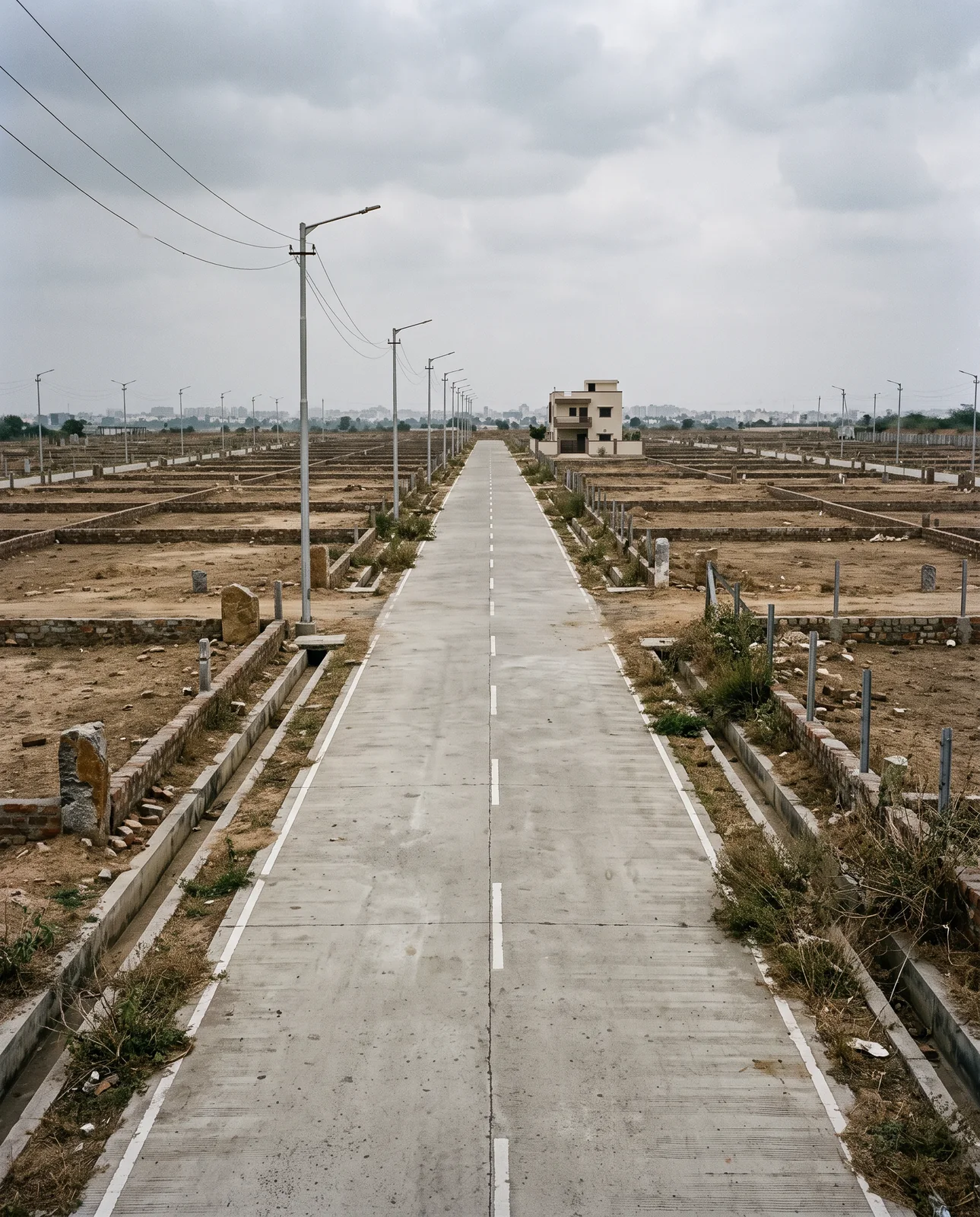 A long concrete road with a dashed centre line runs through a plotted colony of bare earth and low boundary walls, street lights along it and a single finished house at the far end.