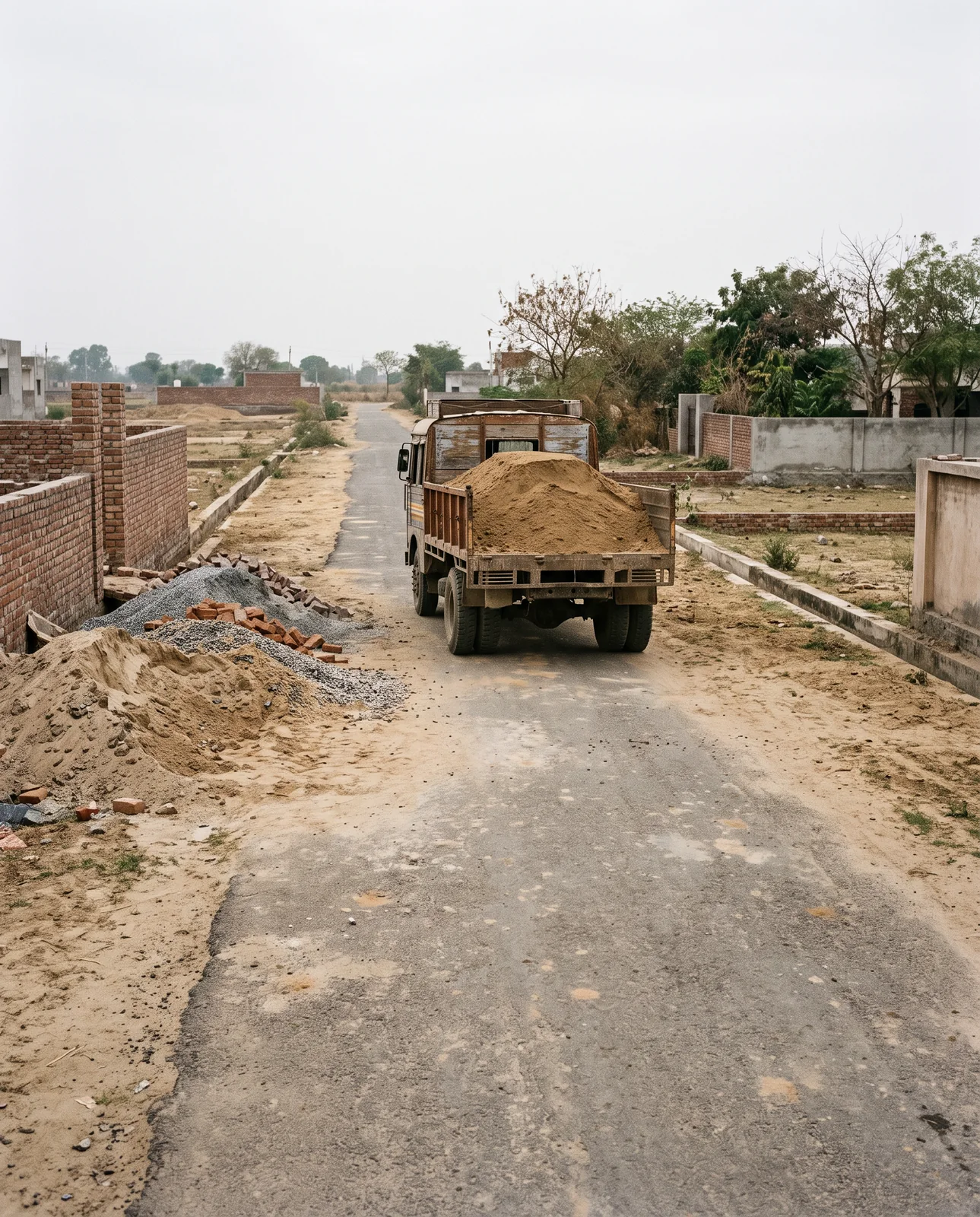 A truck loaded with sand drives down a narrow colony road whose tarmac is hemmed in on one side by heaps of sand, gravel and bricks piled against a half-built brick wall.