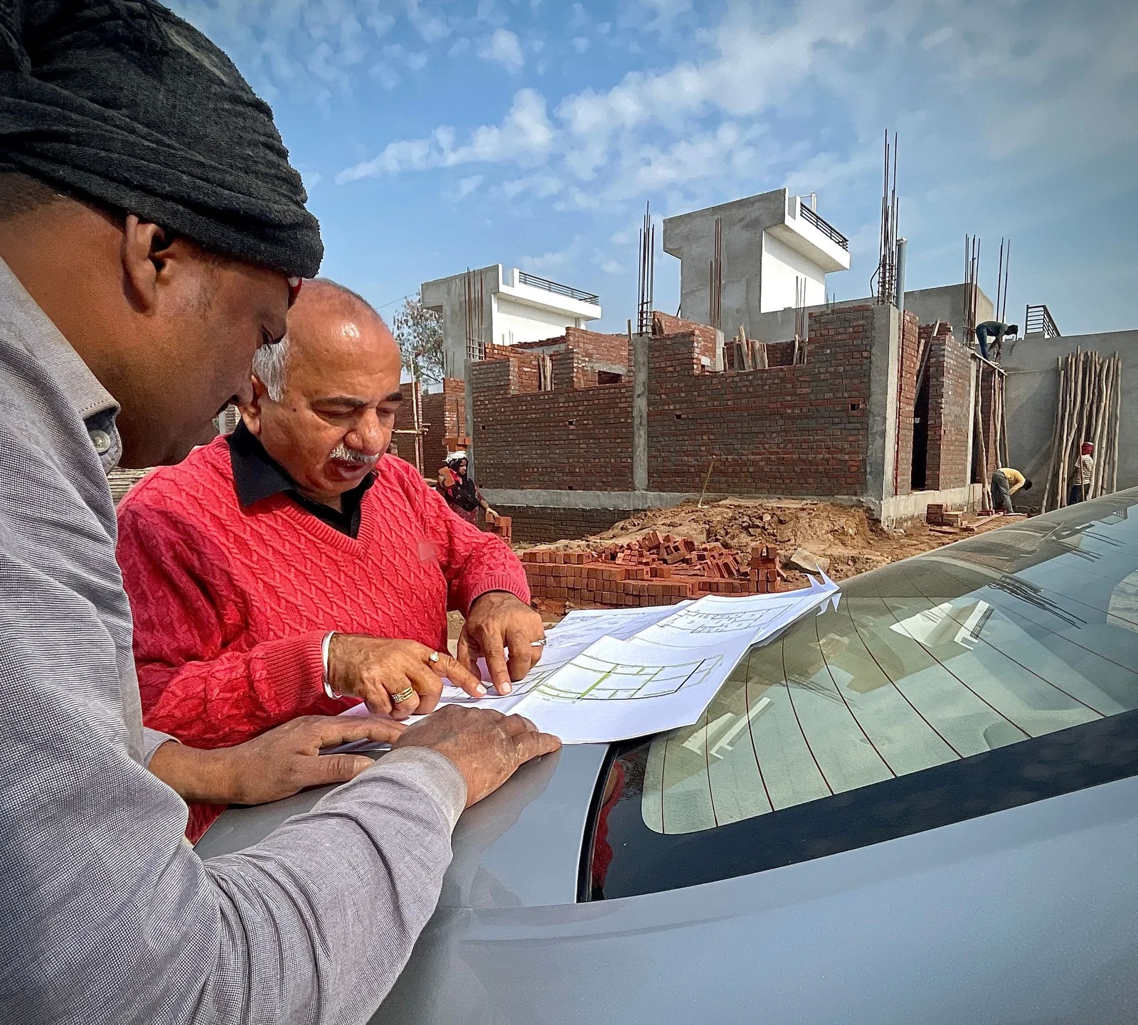 Two men lean over a building plan spread on the bonnet of a silver car, one pointing at the drawing, with half-built brick houses behind them.
