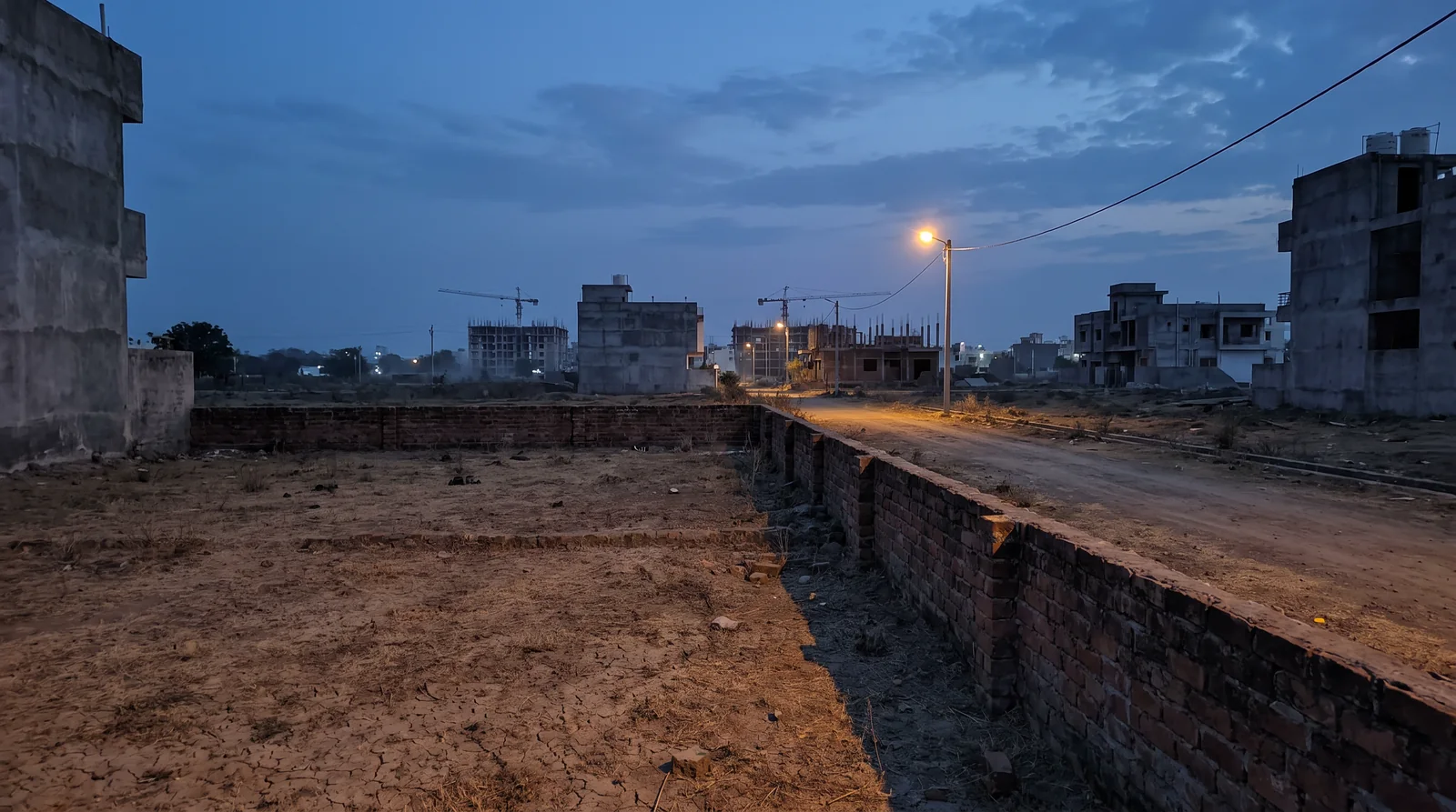 An empty plot enclosed by a low brick wall at dusk, with a streetlight on a dirt road and half-built houses around it.