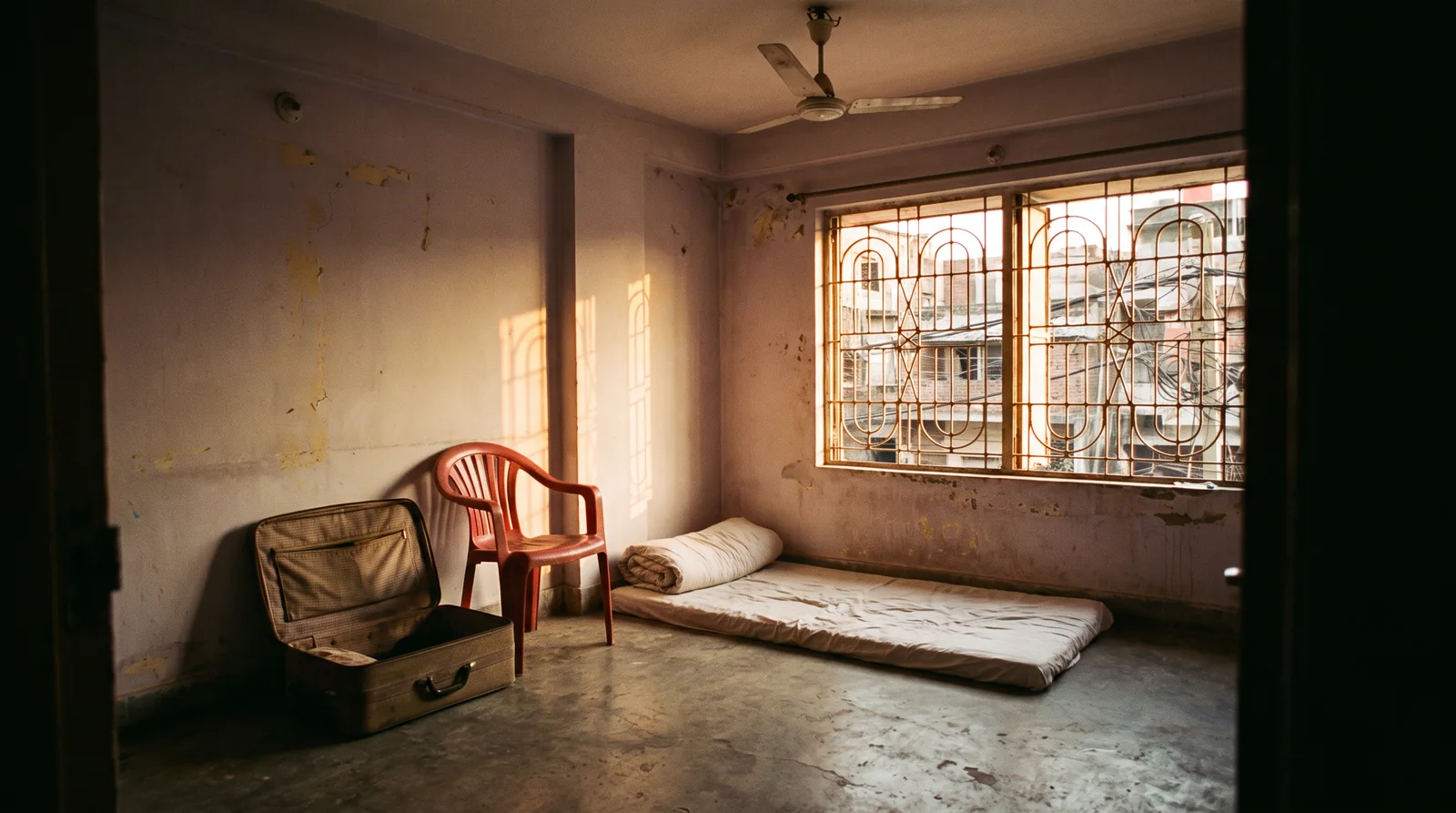 An empty rented room with stained walls, a plastic chair, an open suitcase and a rolled mattress on the floor under a barred window in late sun.