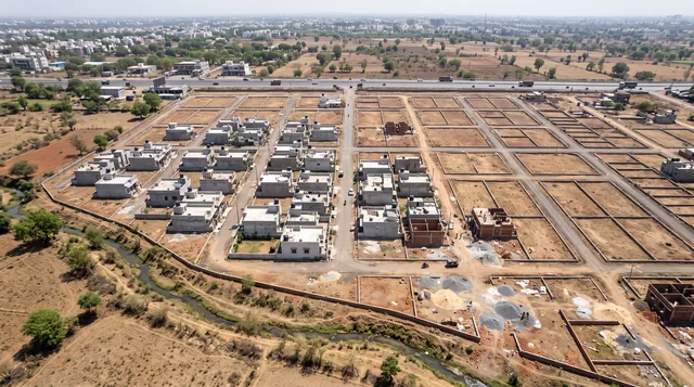 An aerial view of a half-built colony beside a highway, finished grey houses on one side, walled empty plots on the other and a small stream winding past its edge.